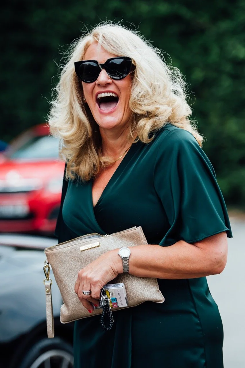A woman with blonde curly hair wearing dark sunglasses and a dark green dress, smiling and holding a beige purse with keys and other items outside with cars and greenery in the background.
