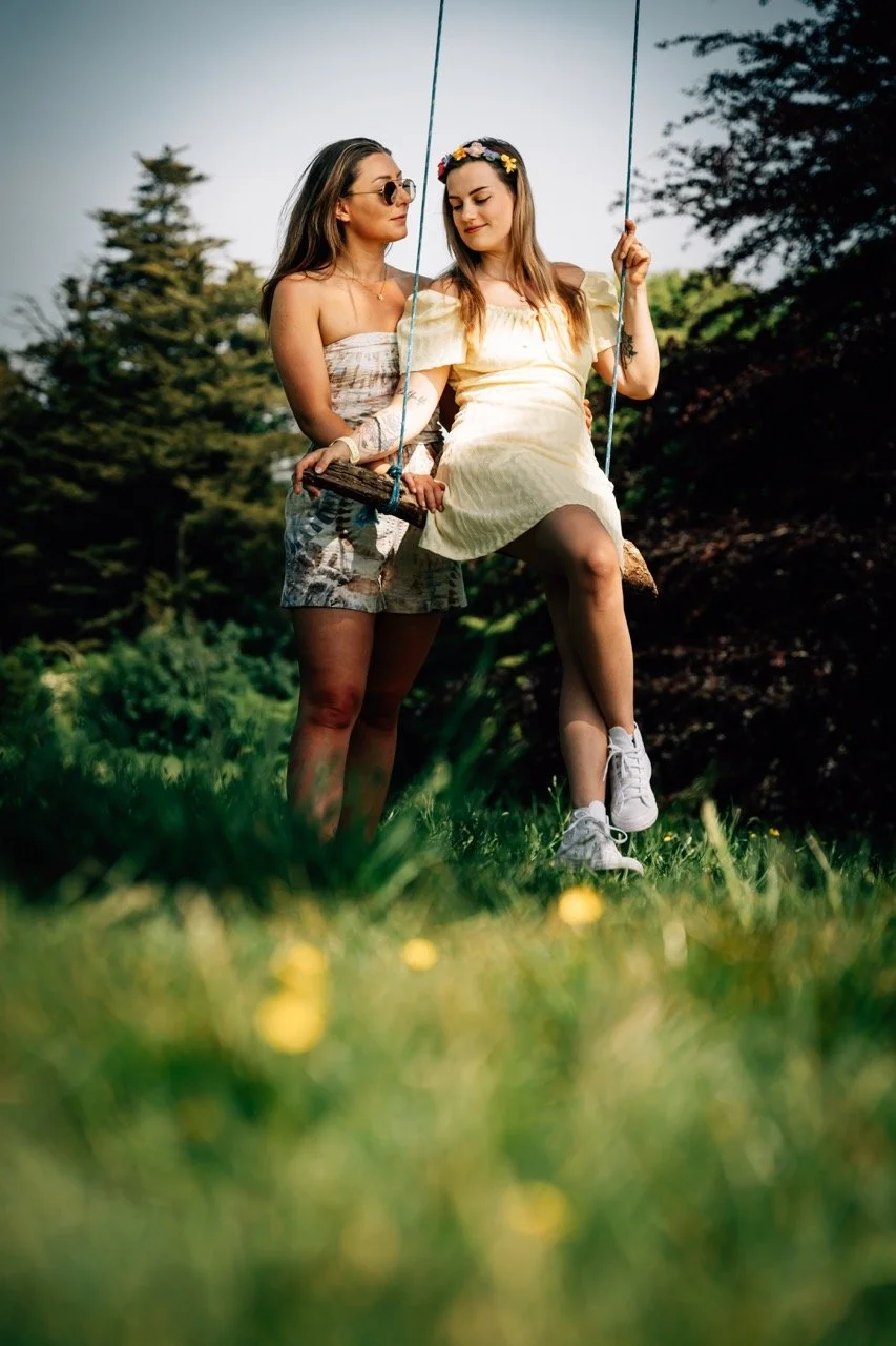 Two young women sitting on a swing outdoors in a grassy area with trees in the background, one wearing sunglasses and the other with a floral headband, enjoying a sunny day.