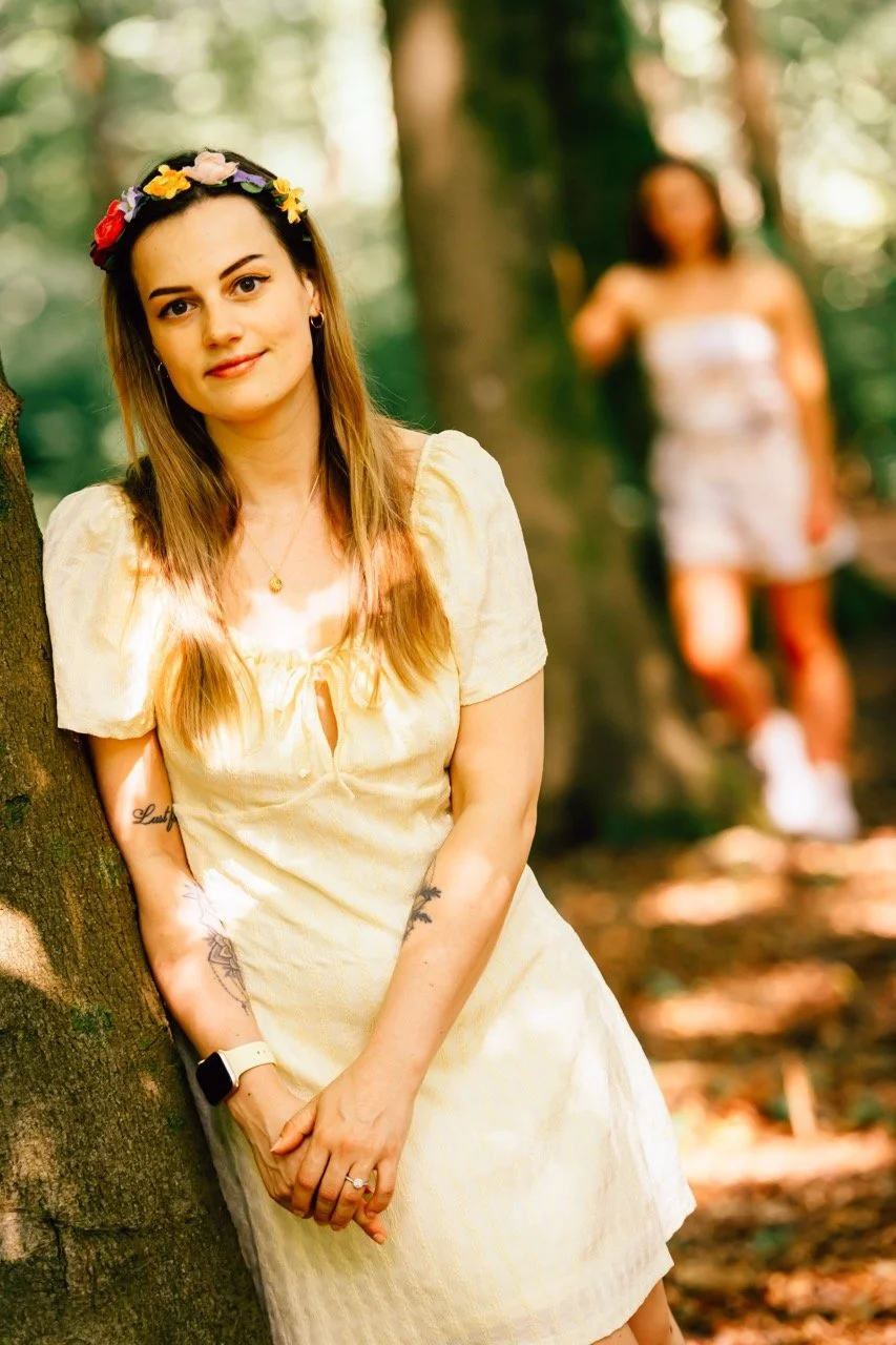 A woman with long light brown hair wearing a floral crown and a pale yellow dress, leaning against a tree in a forest with another woman in the background.