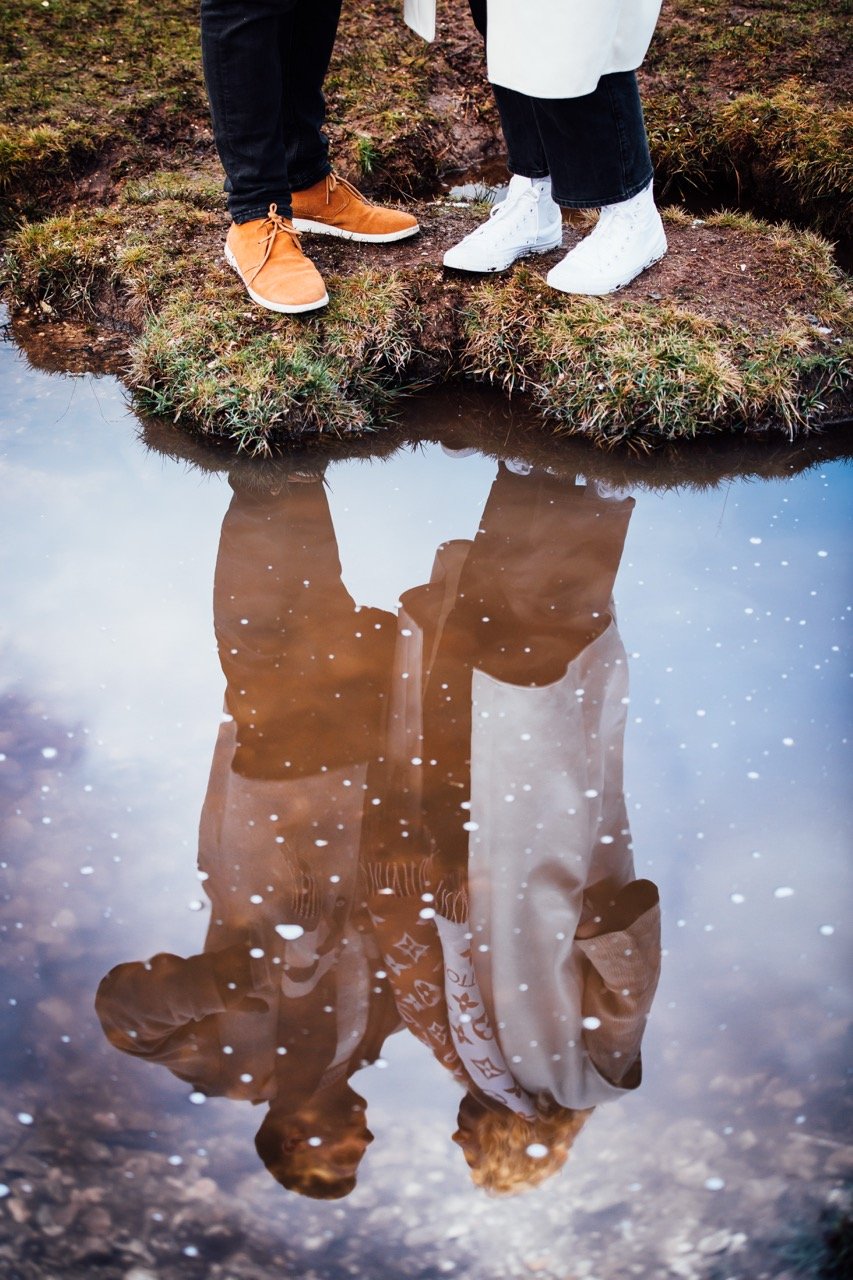 Two people standing on a grassy patch near a small body of water, with their reflection visible in the water, showing their legs and shoes.