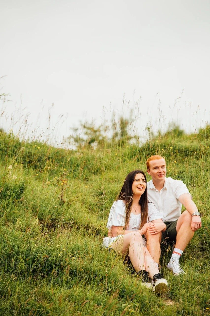 A young woman and a young man sitting on a grassy hillside, smiling and enjoying the outdoors.