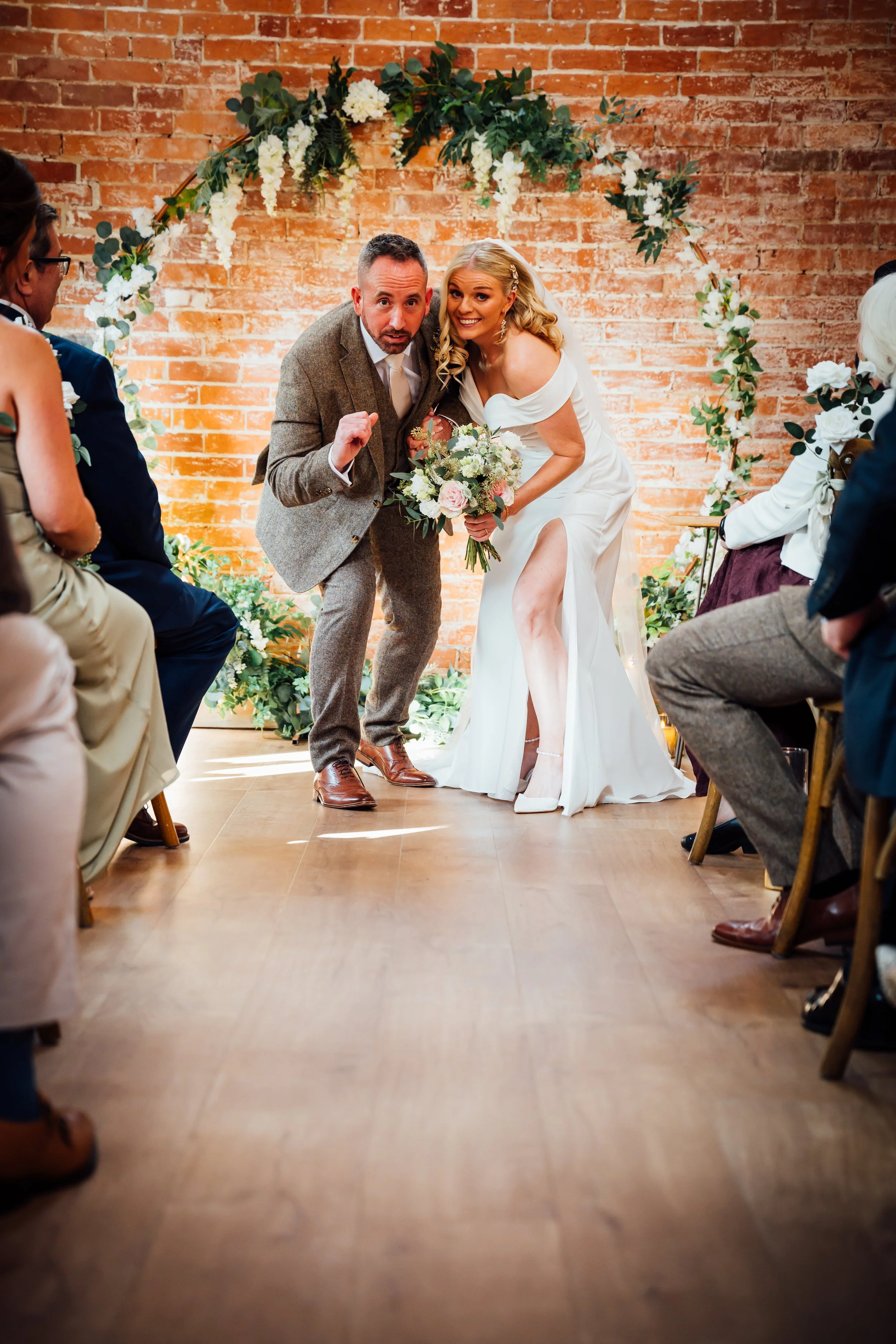 A bride in a white wedding dress with a high slit and off-shoulder design standing next to a man in a gray suit at a wedding ceremony, both leaning in and smiling. The woman holds a bouquet of flowers. They are positioned in front of a brick wall dec