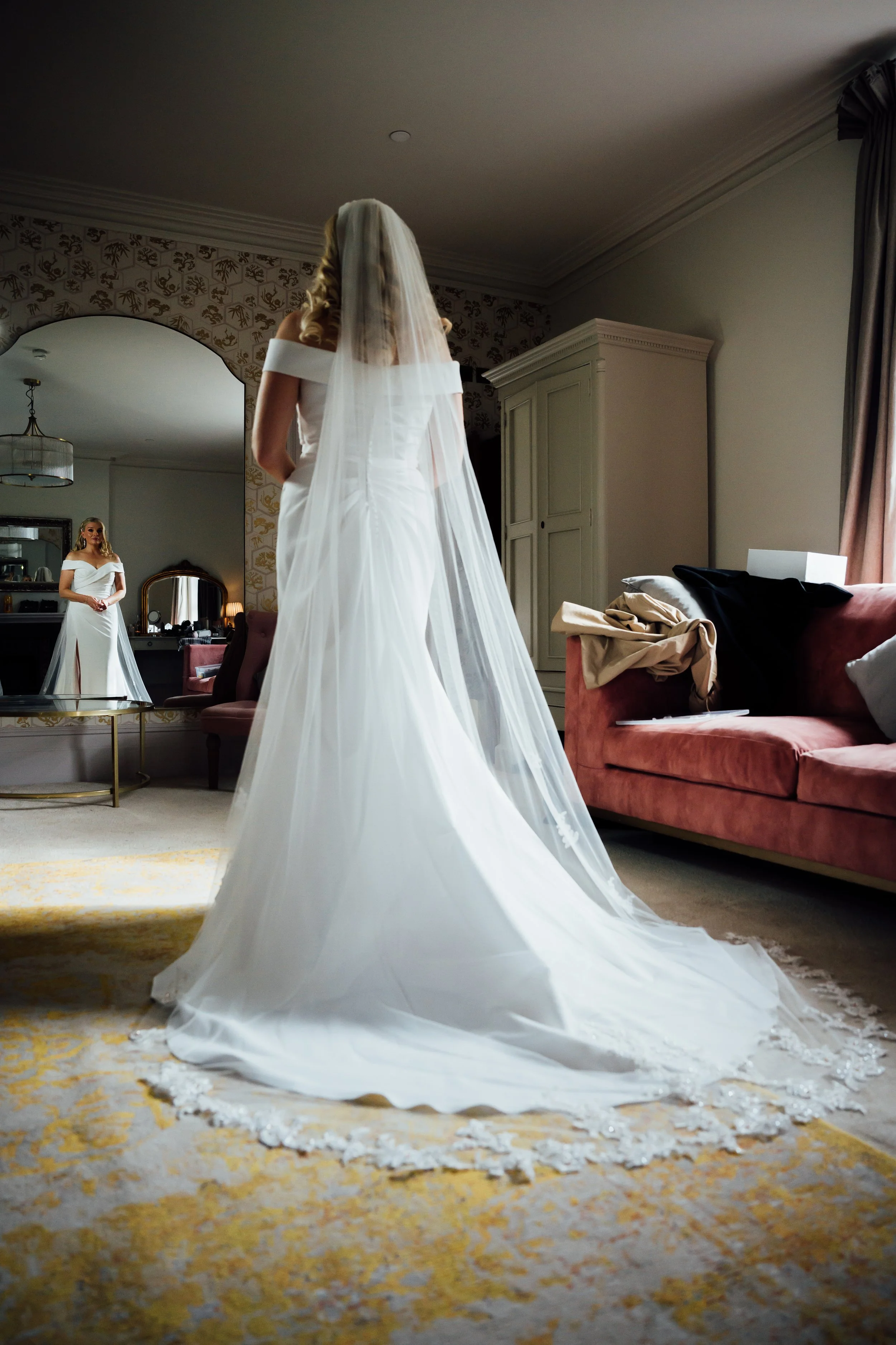 A bride in a wedding gown with a long train and veil, standing in a room looking at her reflection in a mirror.