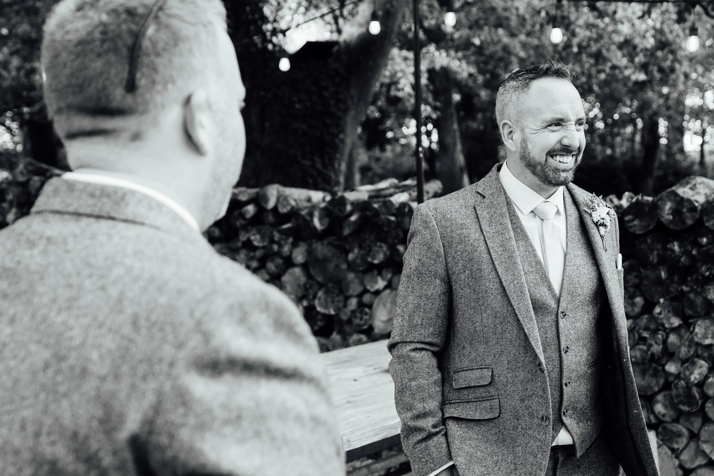 Two men at a wedding, one in the foreground with his back to the camera, and the other in the background smiling, both dressed in suits. The background features trees, stones, and hanging lights.