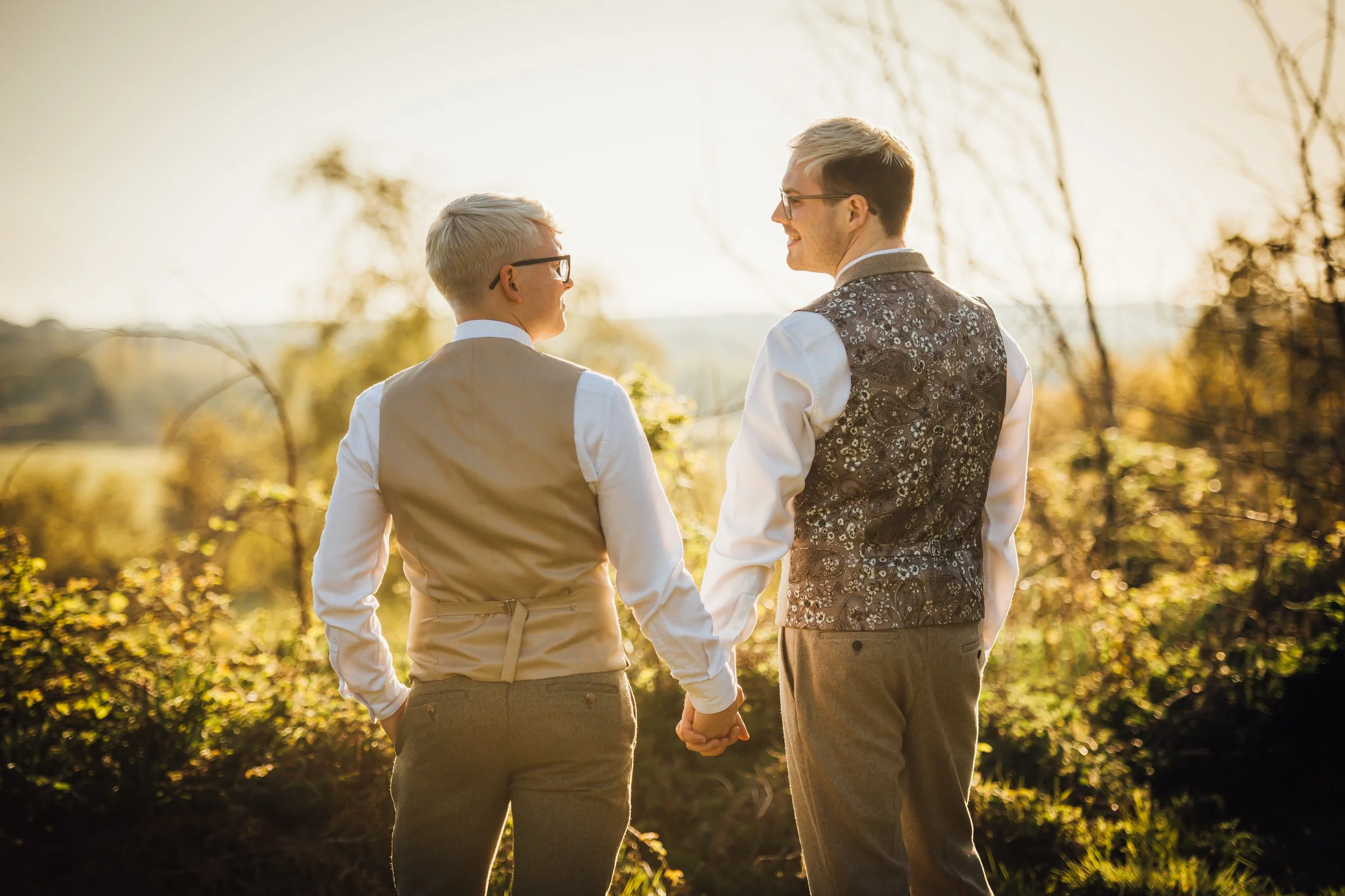 Two people holding hands and smiling, standing outdoors in an autumn landscape with trees and sunlight.