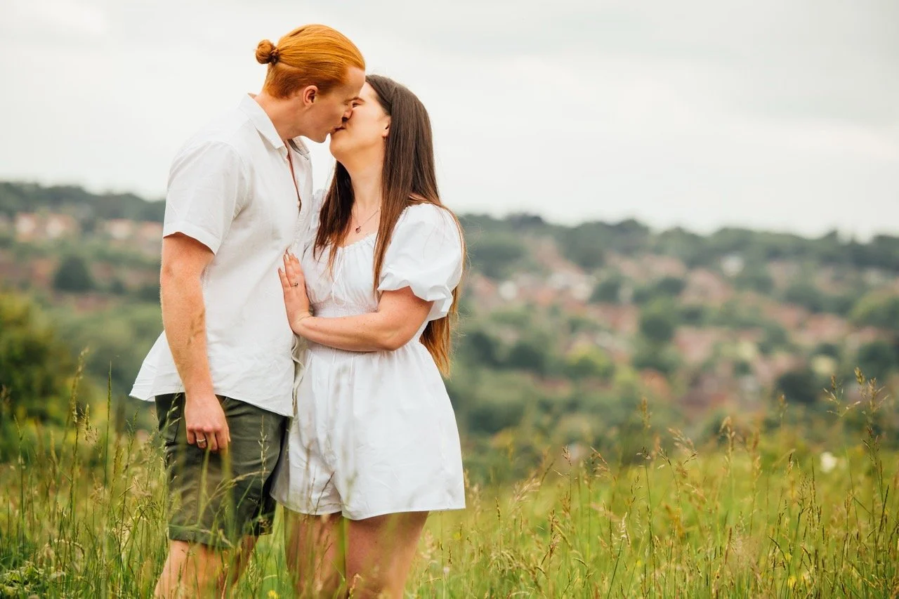 A couple with red hair and long brown hair are kissing in a grassy field with hills and houses in the background.