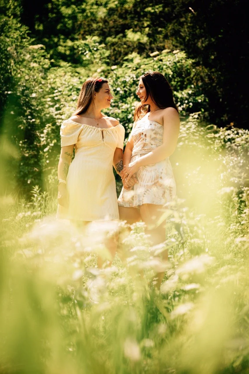 Two women standing in a lush green garden, holding hands and smiling at each other during daytime.