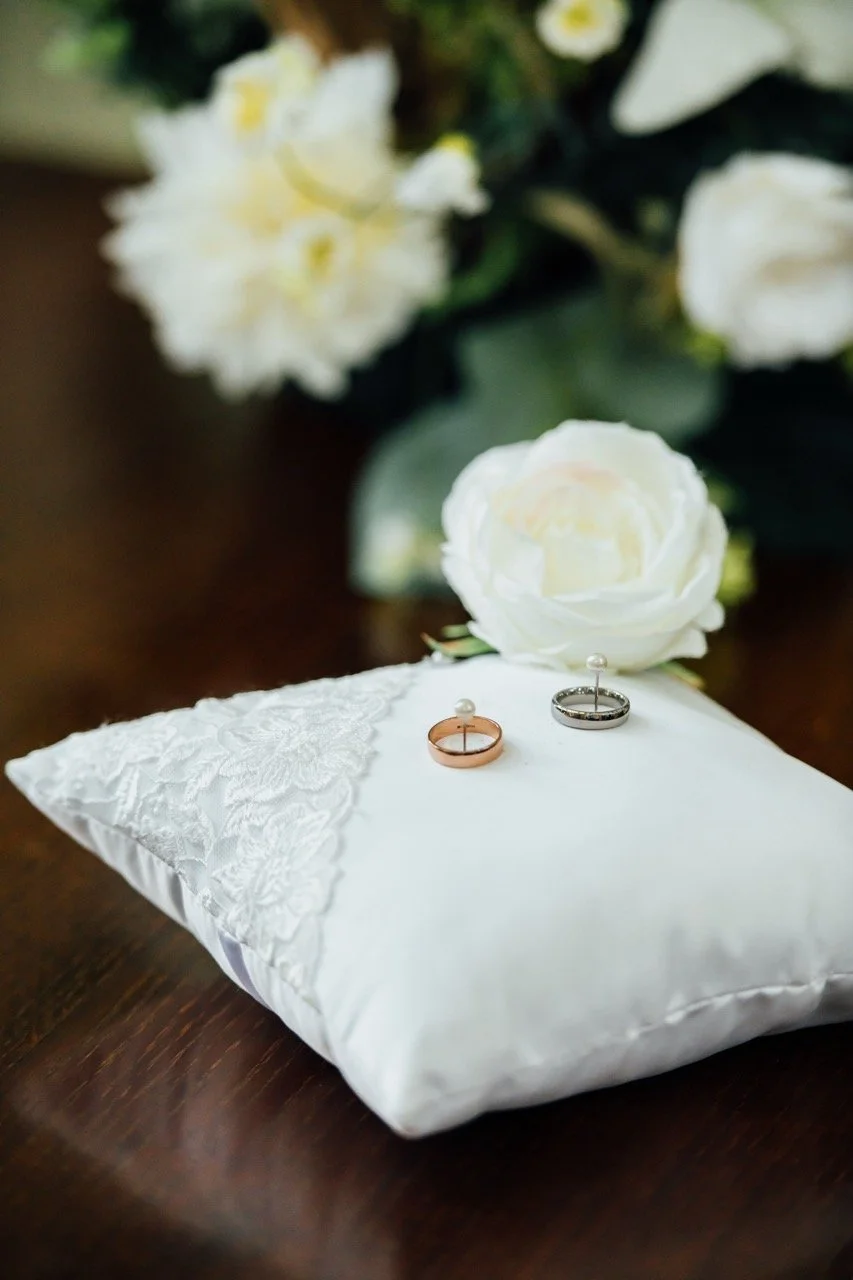 Wedding rings on a white lace pillow, white flowers, and green foliage in the background.