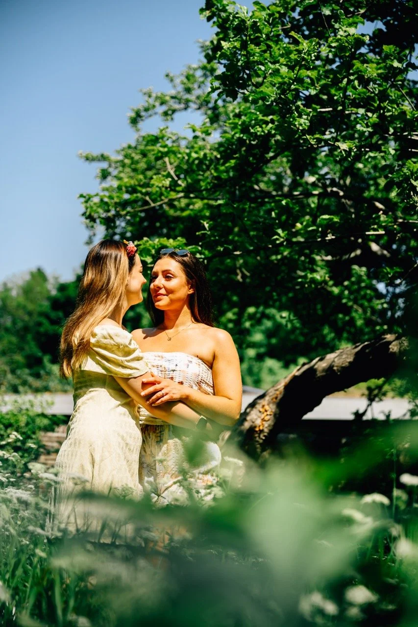 Two women standing outdoors in a lush green park, embracing and smiling at each other on a sunny day.
