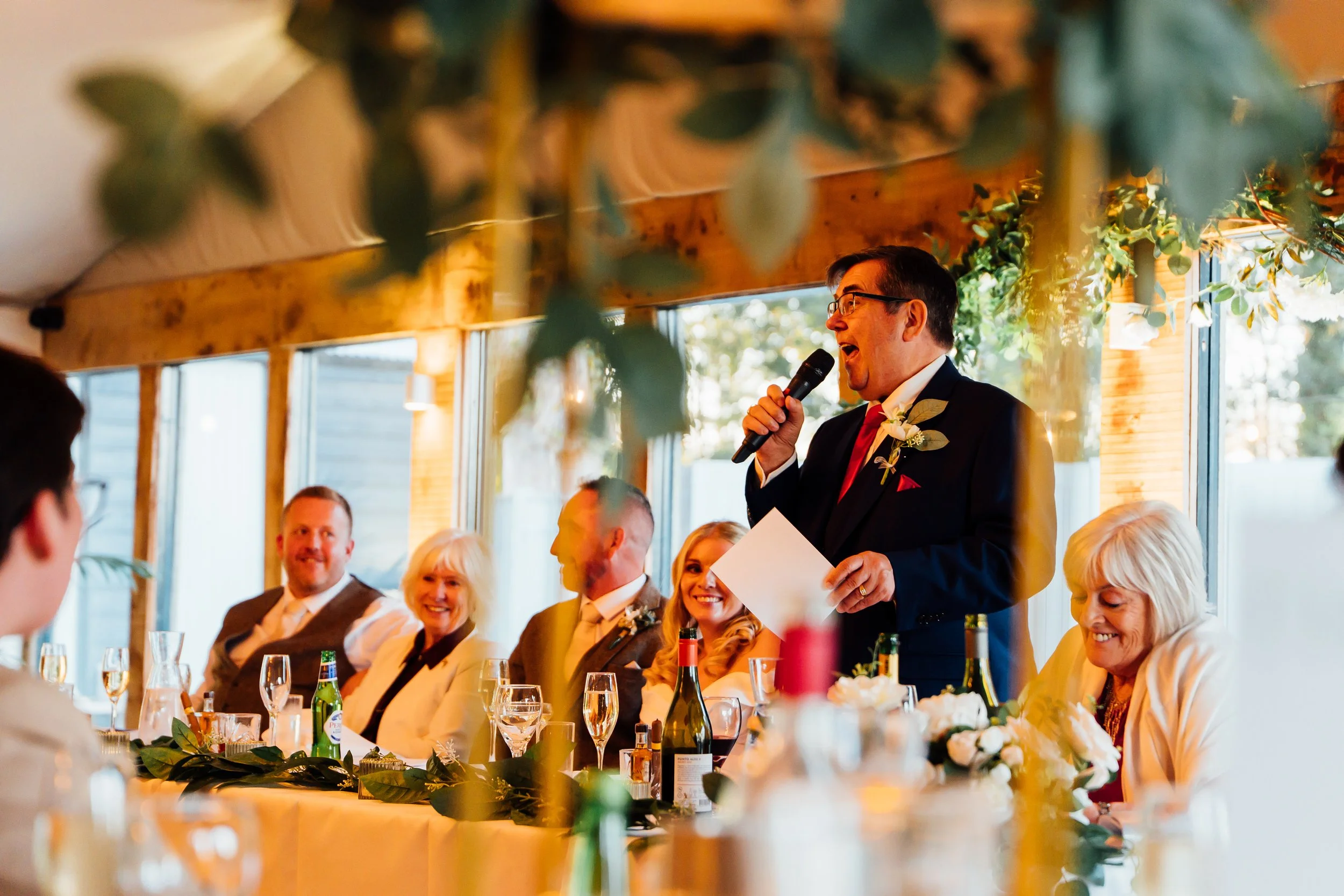 A man in a suit giving a speech at a wedding reception, holding a microphone and notes, seated at a decorated table with guests smiling and listening.