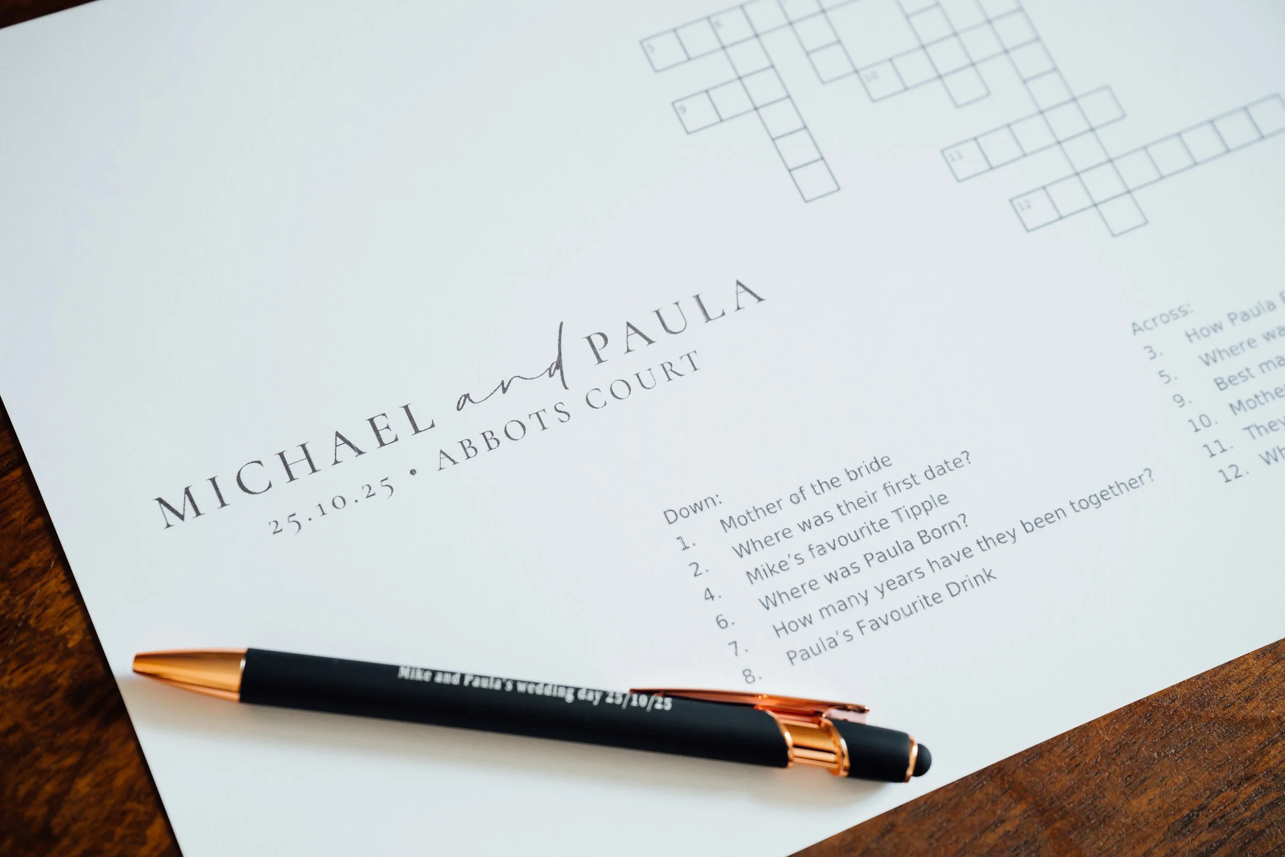 A wedding program with the names Michael and Paula, date October 25, 2023, and location Abbotts Court, placed on a wooden surface. A black and rose gold pen rests on the program.