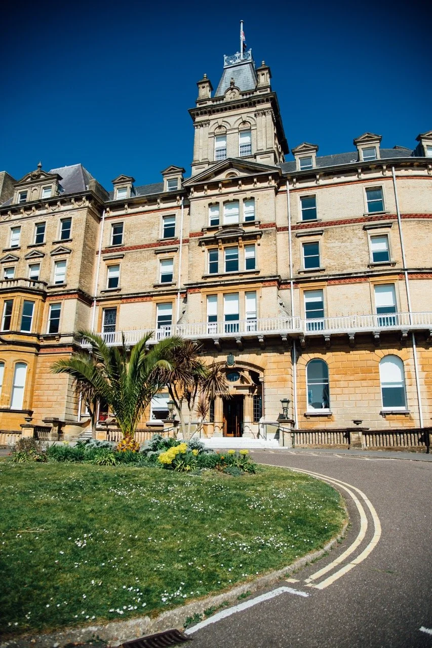A historic building with a tower, beige brick exterior, and a small landscaped garden with palm trees in front, under a clear blue sky.