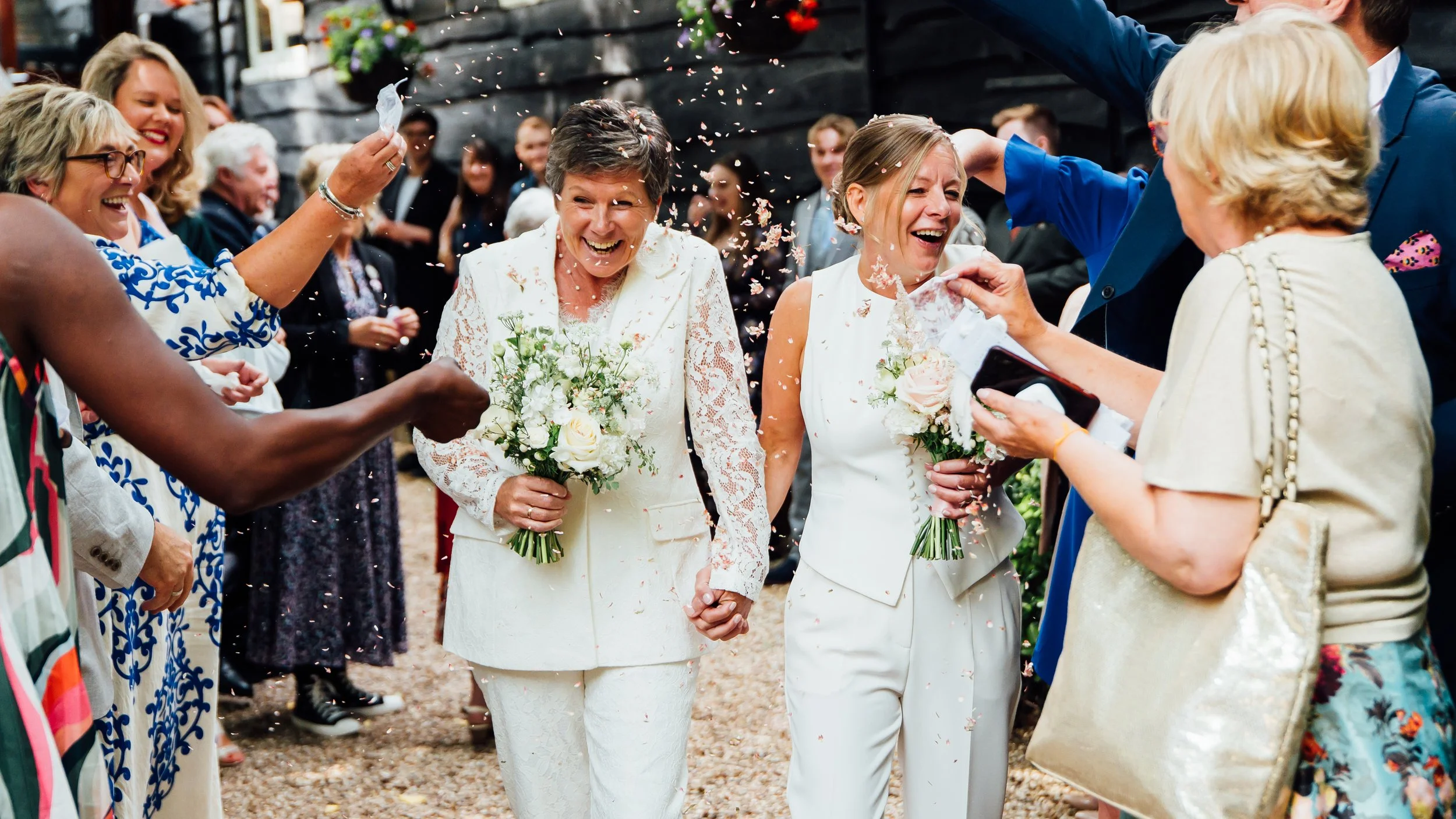 Two women in white outfits holding bouquets, smiling and walking hand in hand at a wedding celebration, surrounded by guests throwing flower confetti.