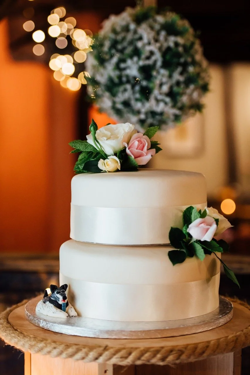 Two-tiered white wedding cake decorated with pink and white flowers and green leaves, with a small dog figurine at the base, placed on a wooden stand with a braided rope trim. Blurred holiday lights and a snow-dusted Christmas tree are in the backgro