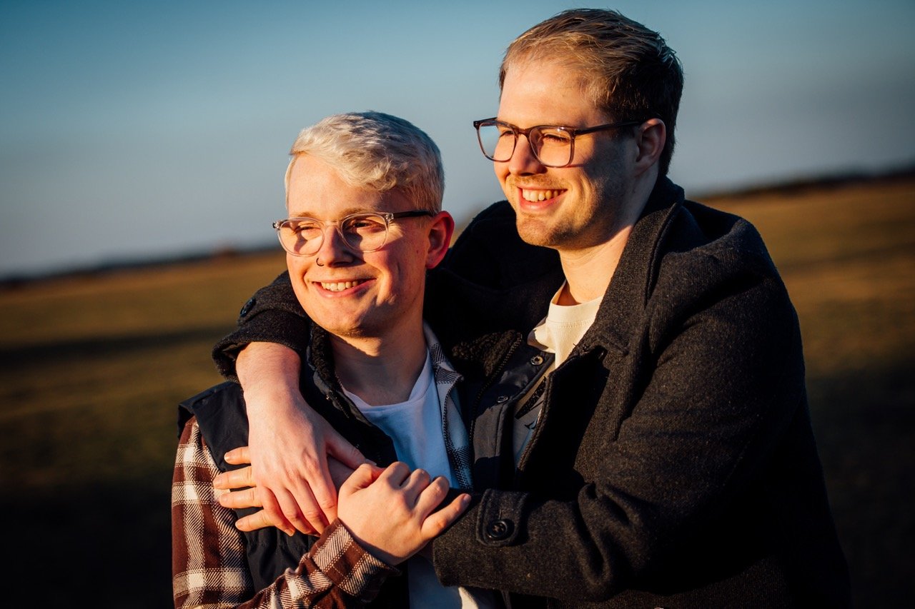 Two young men smiling and embracing outdoors during sunset, with a blurred natural landscape in the background.