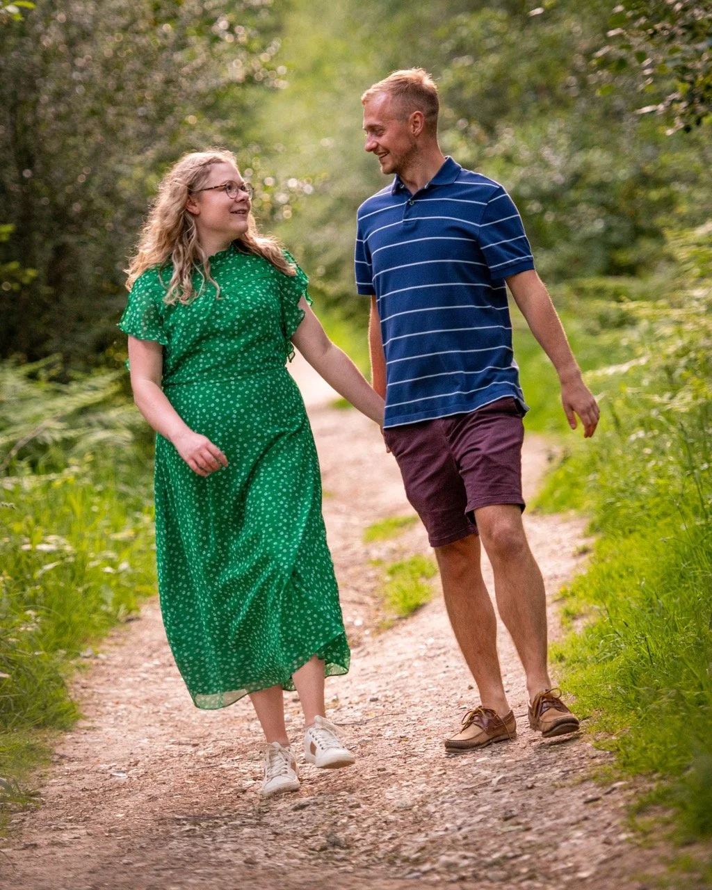 A man and a girl walking hand in hand on a dirt trail surrounded by green trees and grass, smiling at each other.