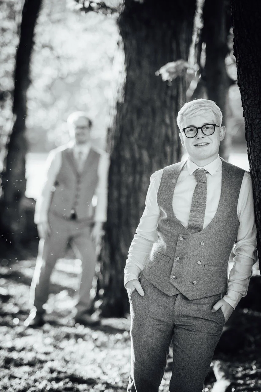 Young man with glasses and light-colored hair standing outdoors by a tree, smiling, with another man blurred in the background. Both are dressed in formal attire, suggesting a vintage or retro style.
