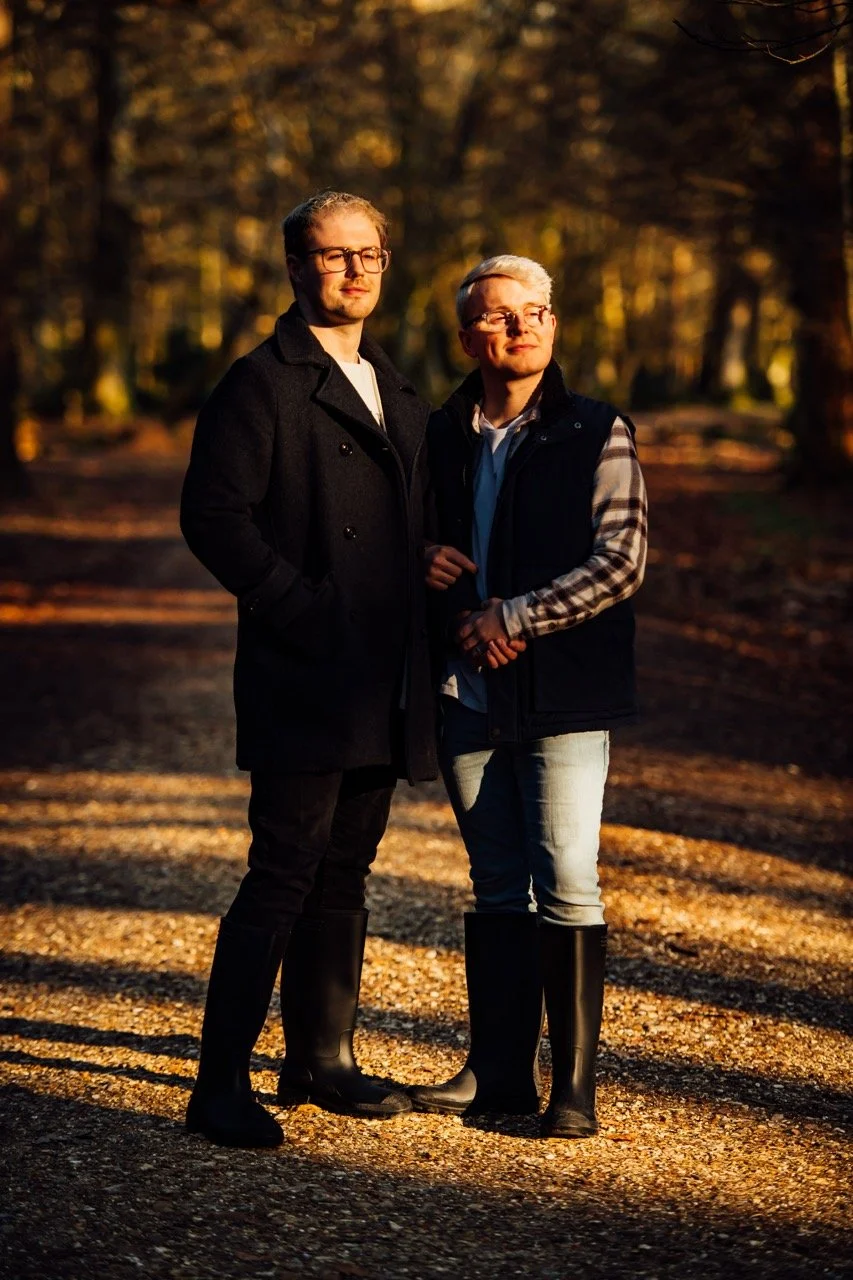 Two men standing outdoors on a wooded path during sunset, both wearing rain boots, warm jackets, and glasses, with trees and falling leaves in the background.