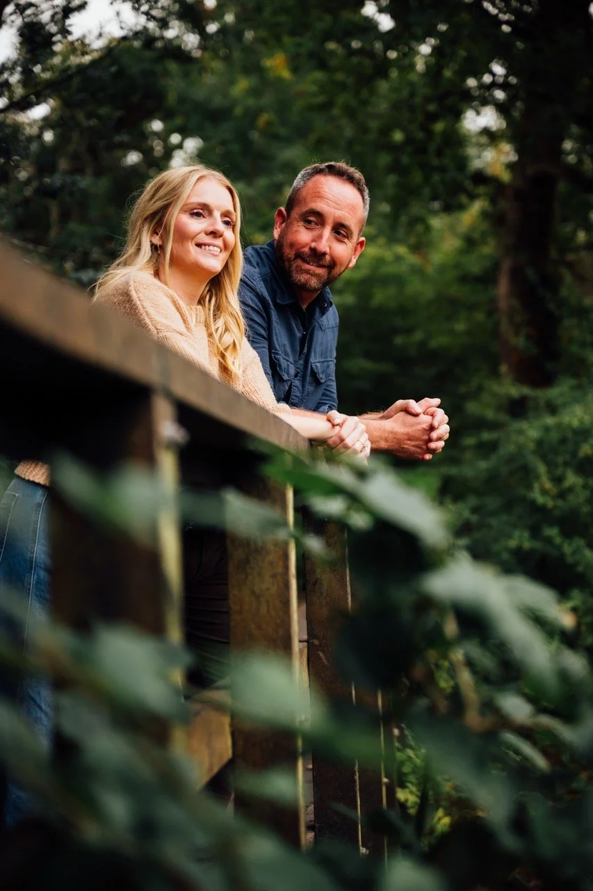A man and woman leaning on a wooden railing outdoors, surrounded by green trees and foliage, smiling and enjoying nature.