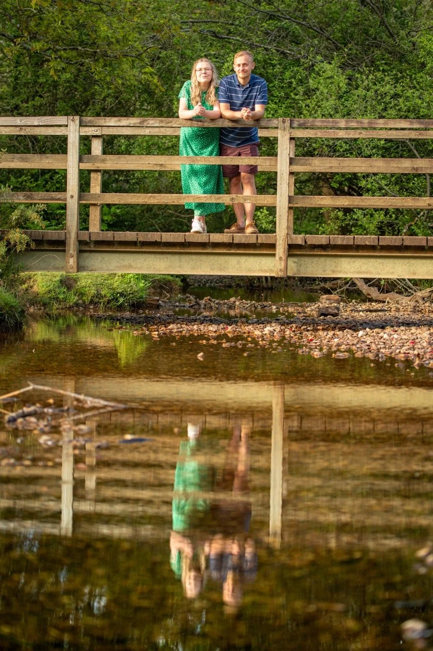 Two people stands on a wooden bridge over a creek, with trees in the background. They are posing for a photo and looking at the camera; the person on the left is in a green dress, and the person on the right is in a striped shirt and shorts. Their re