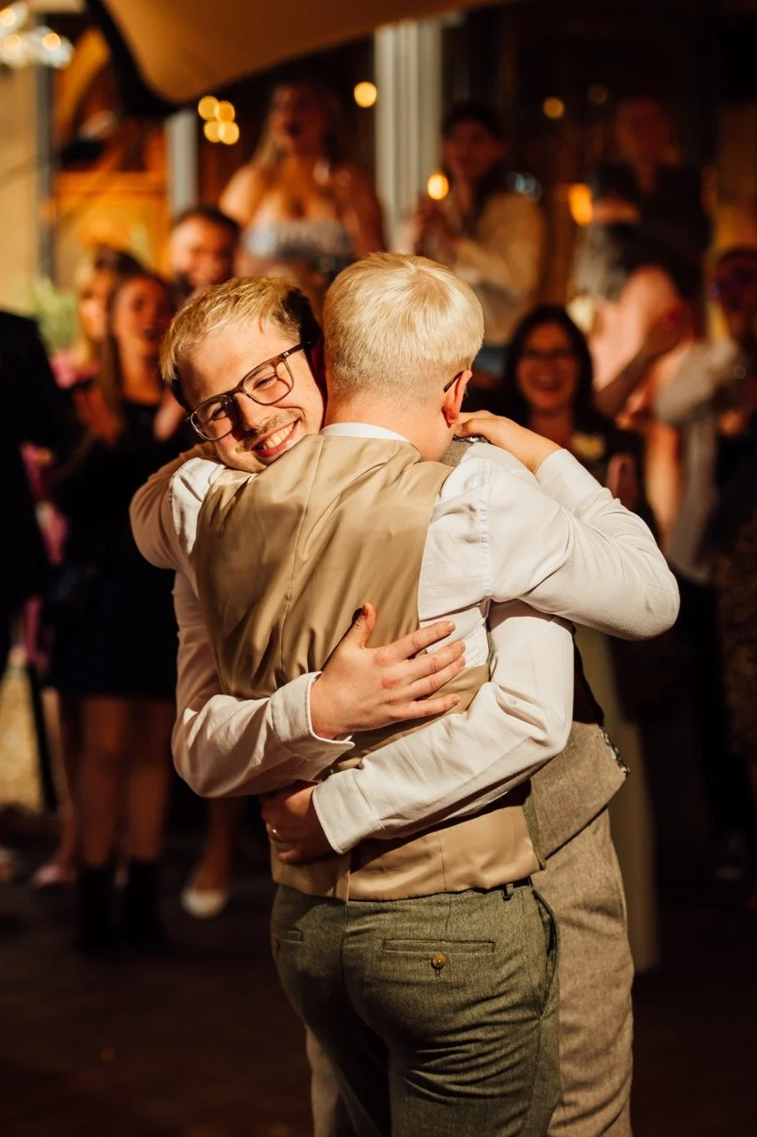 Two young men are hugging at a lively party with smiling and laughing guests in the background.