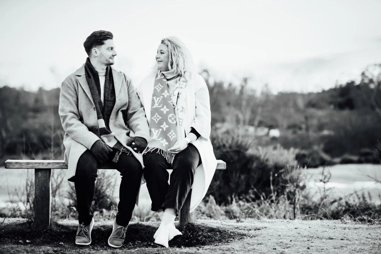 A man and woman sitting on a bench outdoors, looking at each other, with a blurred natural landscape background in black and white.