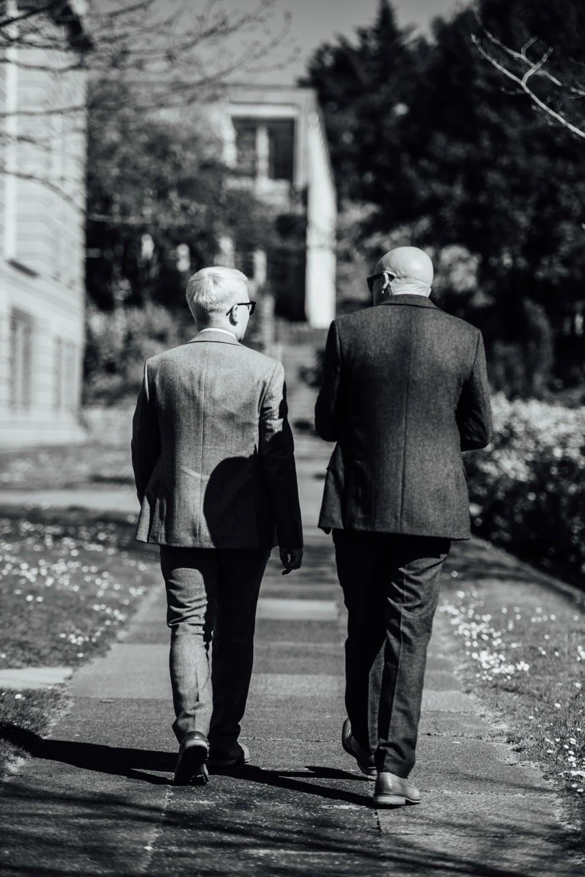 Two men in suits walking down a sidewalk on a sunny day, viewed from behind, with trees and houses in the background.