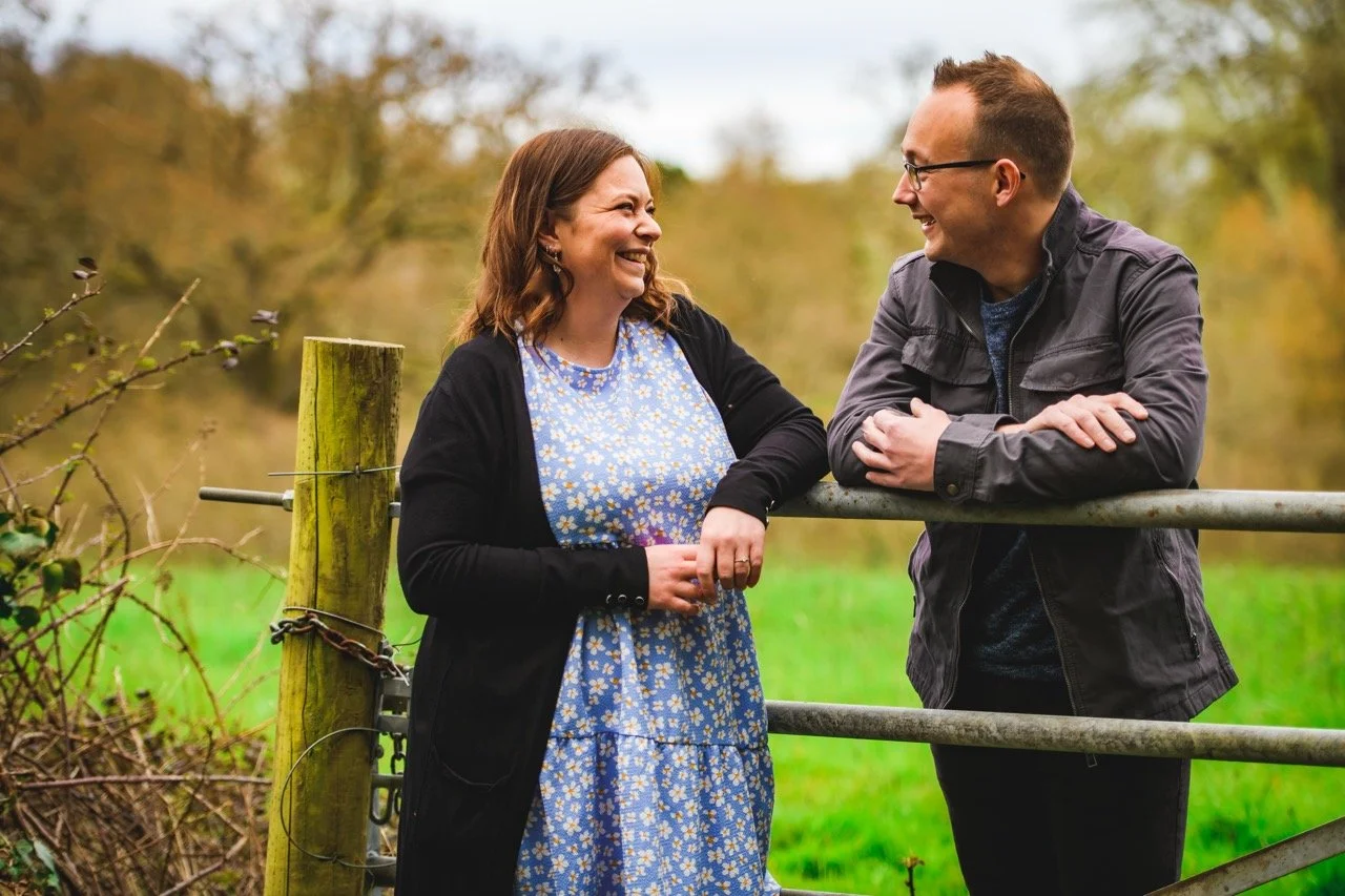 A man and woman smiling and talking outdoors near a metal gate in a rural setting with trees and grass in the background.