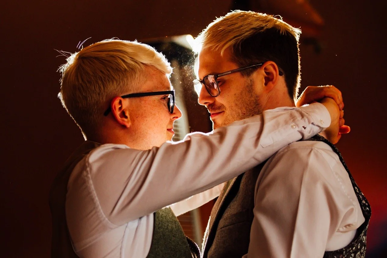 Two men with glasses dancing closely and smiling at each other, illuminated by warm light.