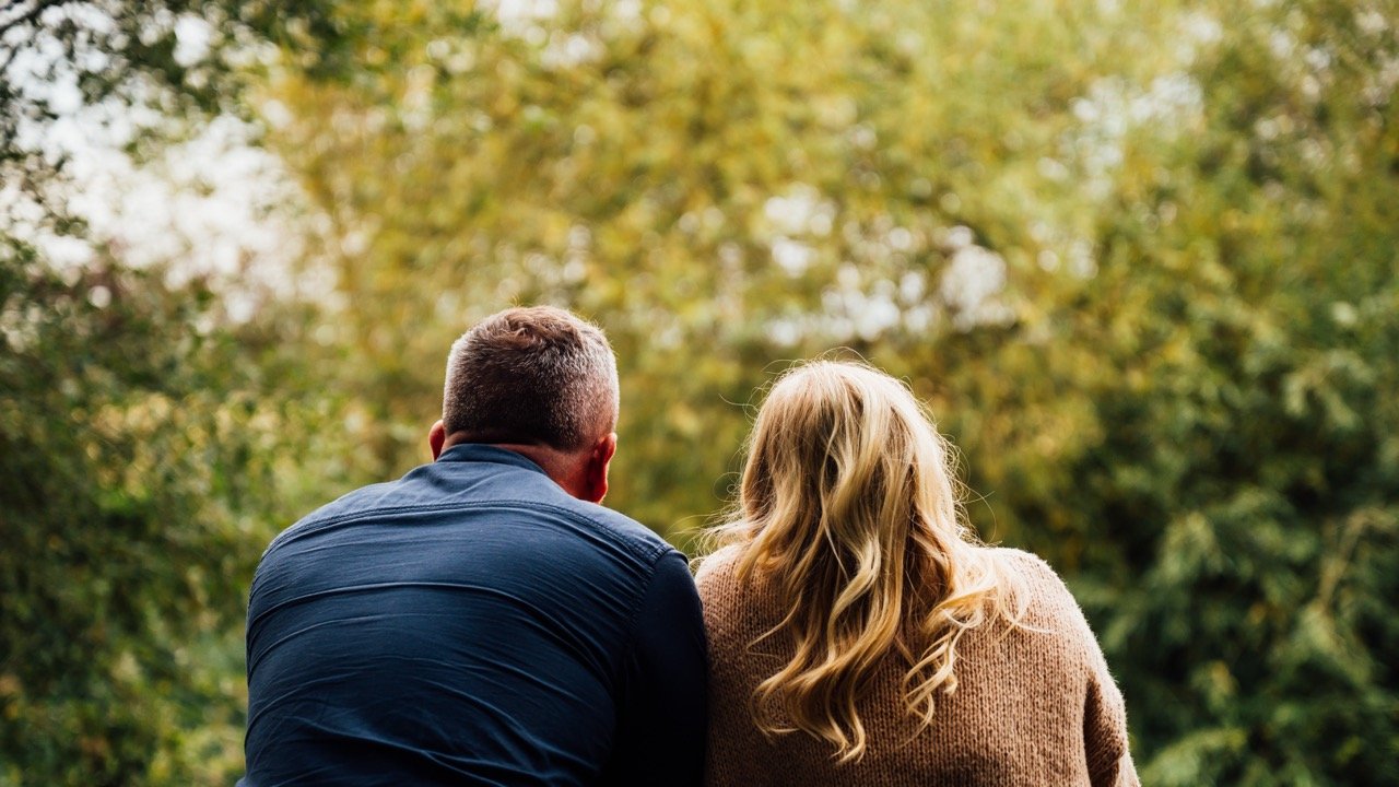 A man and a woman sitting closely outdoors, with trees and autumn foliage in the background.