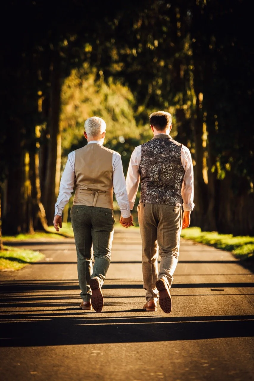 Two people, dressed in vintage-style clothing, walk hand-in-hand down a sunlit path lined with trees in the evening or late afternoon.