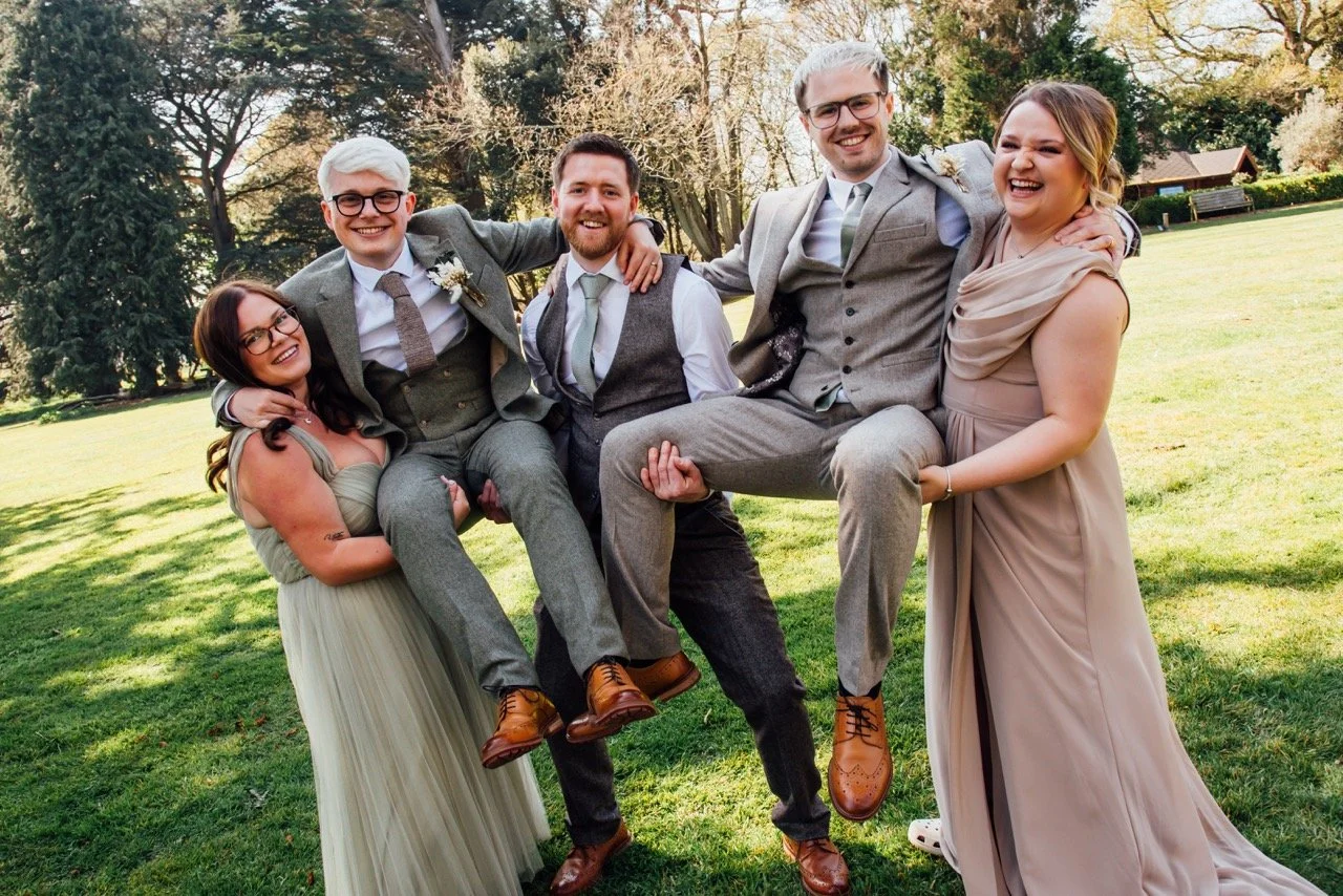 Group of six people smiling and lifting each other in a park with trees in the background during daytime. Three women in dresses and three men in suits.