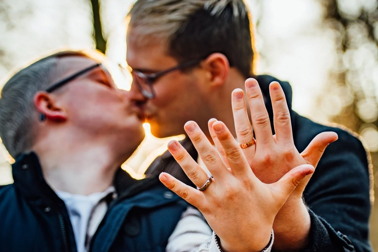 A couple kissing outdoors with sunlight shining through trees in the background; focus on their hands with wedding rings.