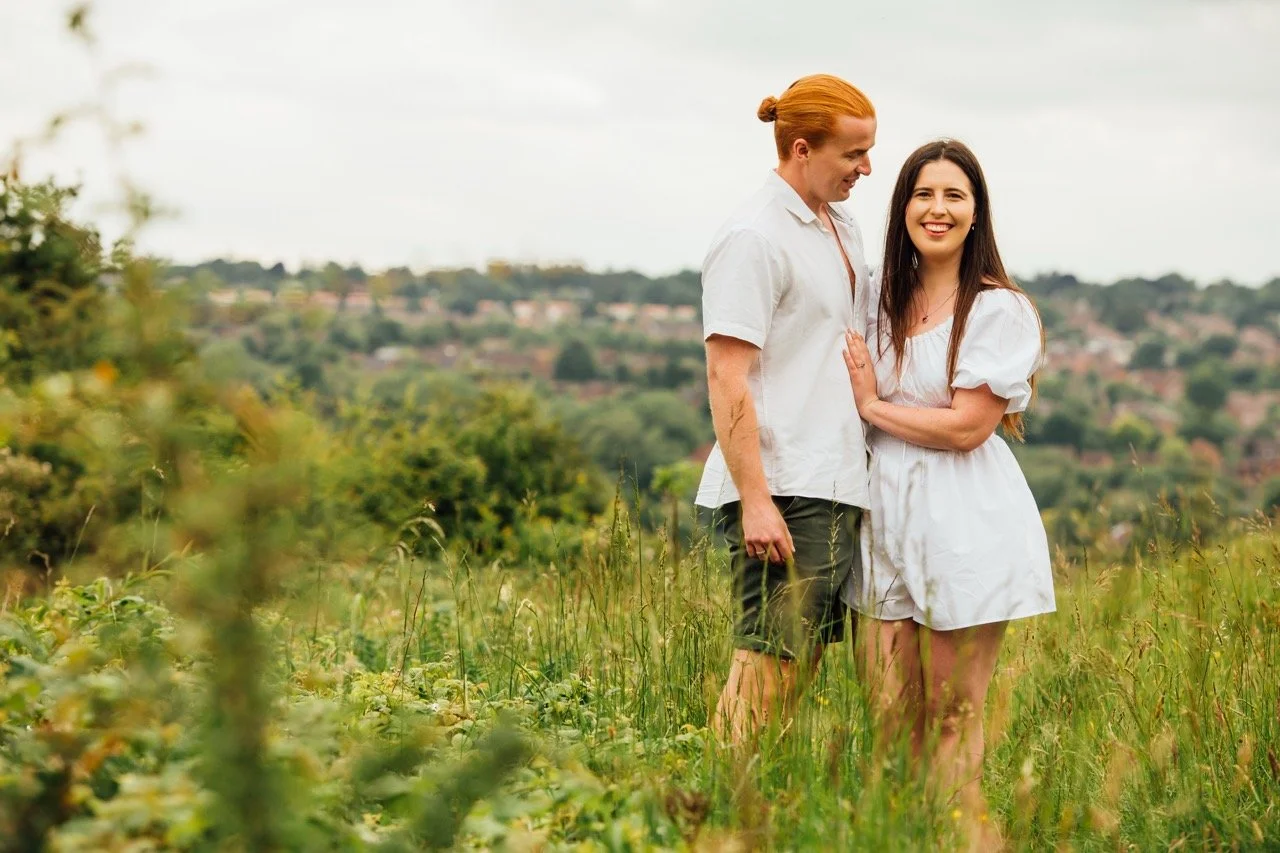 A couple standing together in a grassy field, smiling, with a hilly landscape and houses in the background.