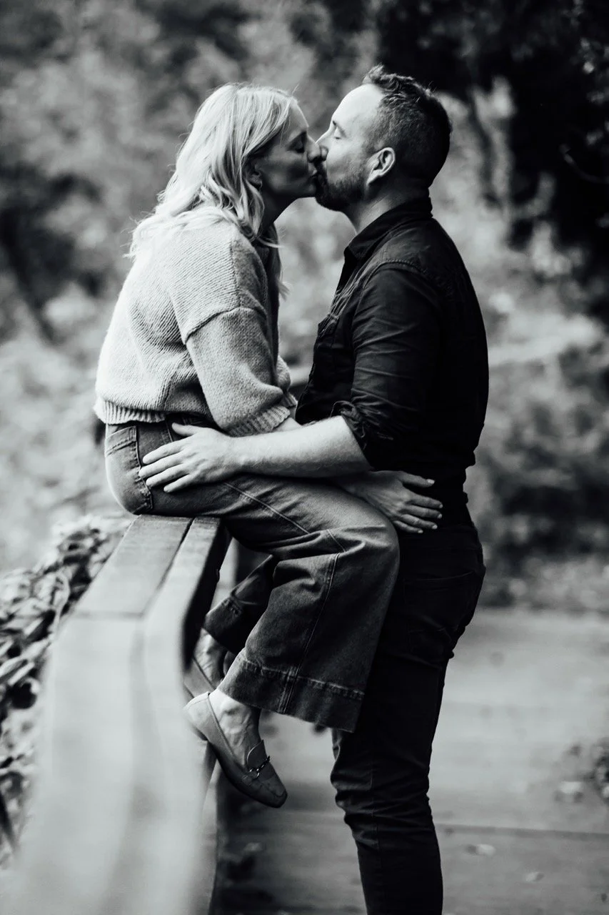 A black-and-white photo of a couple kissing outdoors. The woman is sitting on a park bench, and the man is standing, holding her. Trees are blurred in the background.