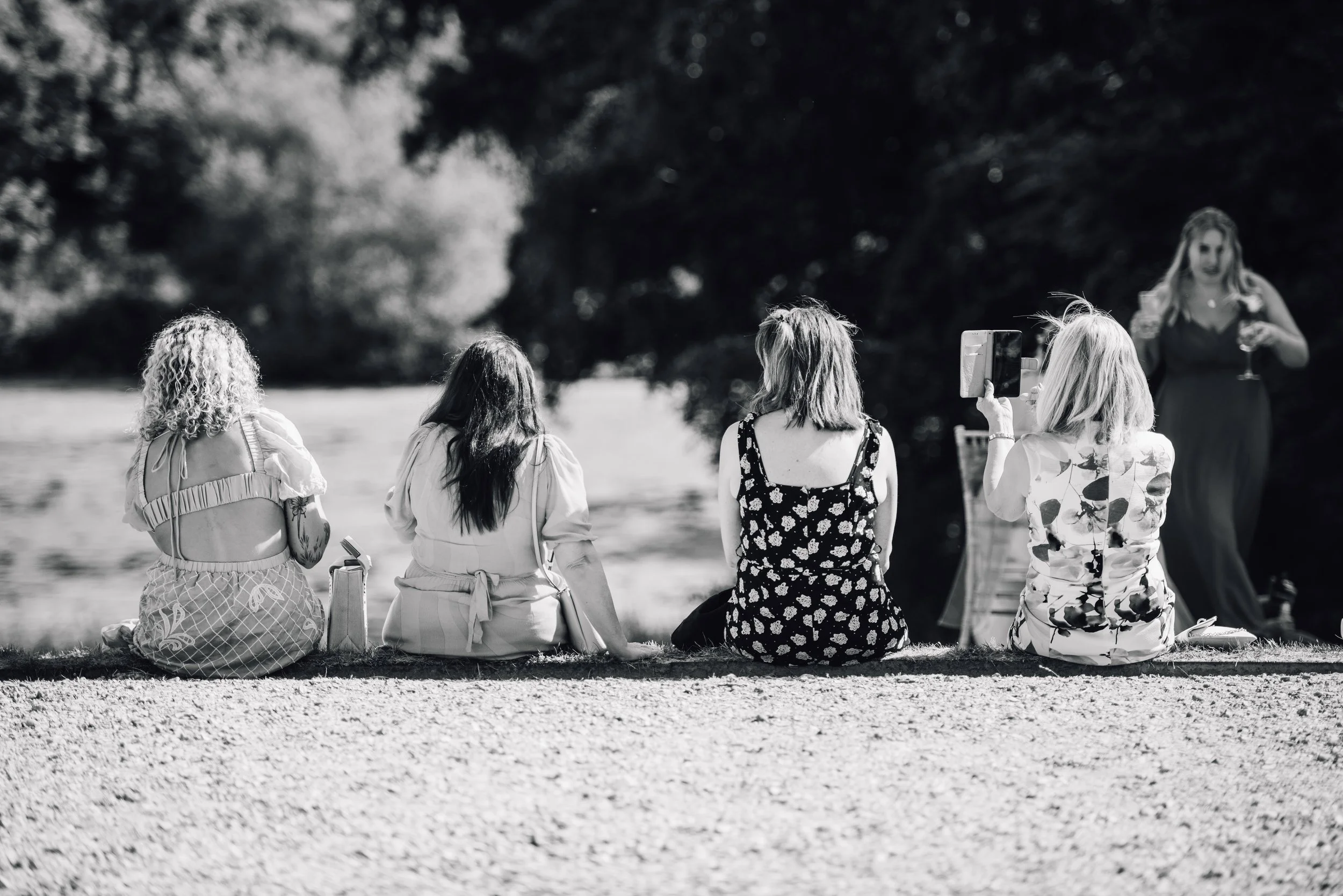 Four women sitting on the grass, facing a lake or river, with a woman standing nearby holding a glass of wine, surrounded by trees.