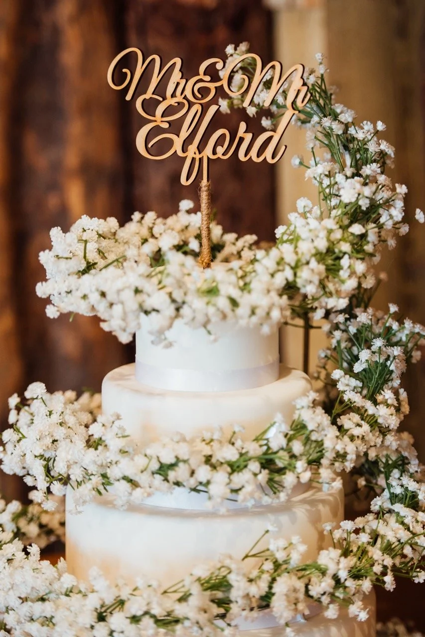 A three-tier white wedding cake decorated with white flowers and a gold cake topper that reads 'Mom of the Bride'.