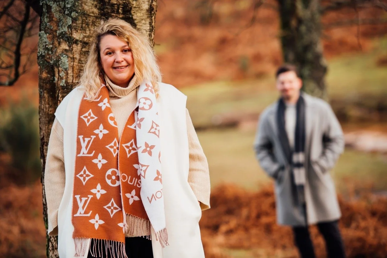 A woman with blonde hair smiling in autumn outdoors, wearing a beige sweater and a Louis Vuitton scarf. A man stands in the background near a tree, dressed in a grey coat and black scarf, blurred out, in a park with fall foliage.