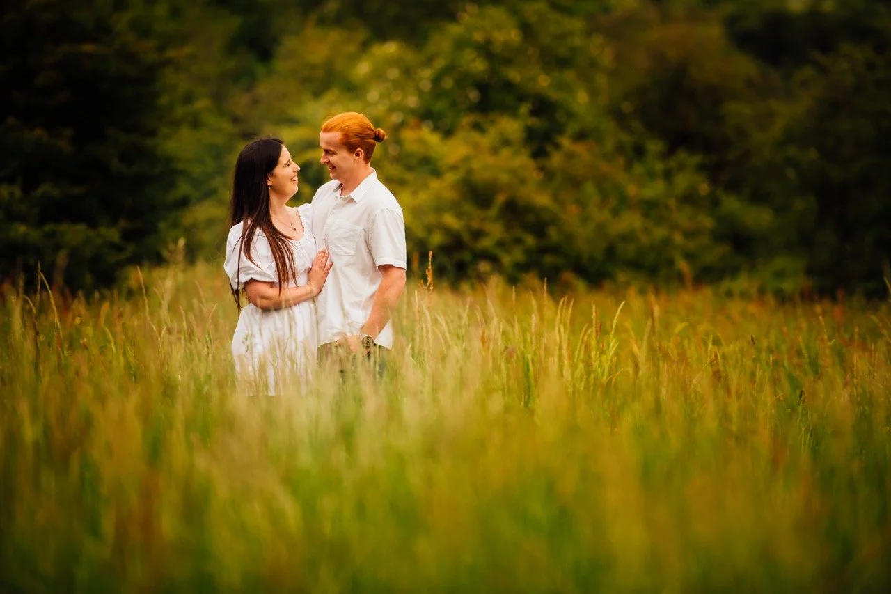 A couple standing in a grassy field, smiling and looking at each other with trees in the background during daytime.