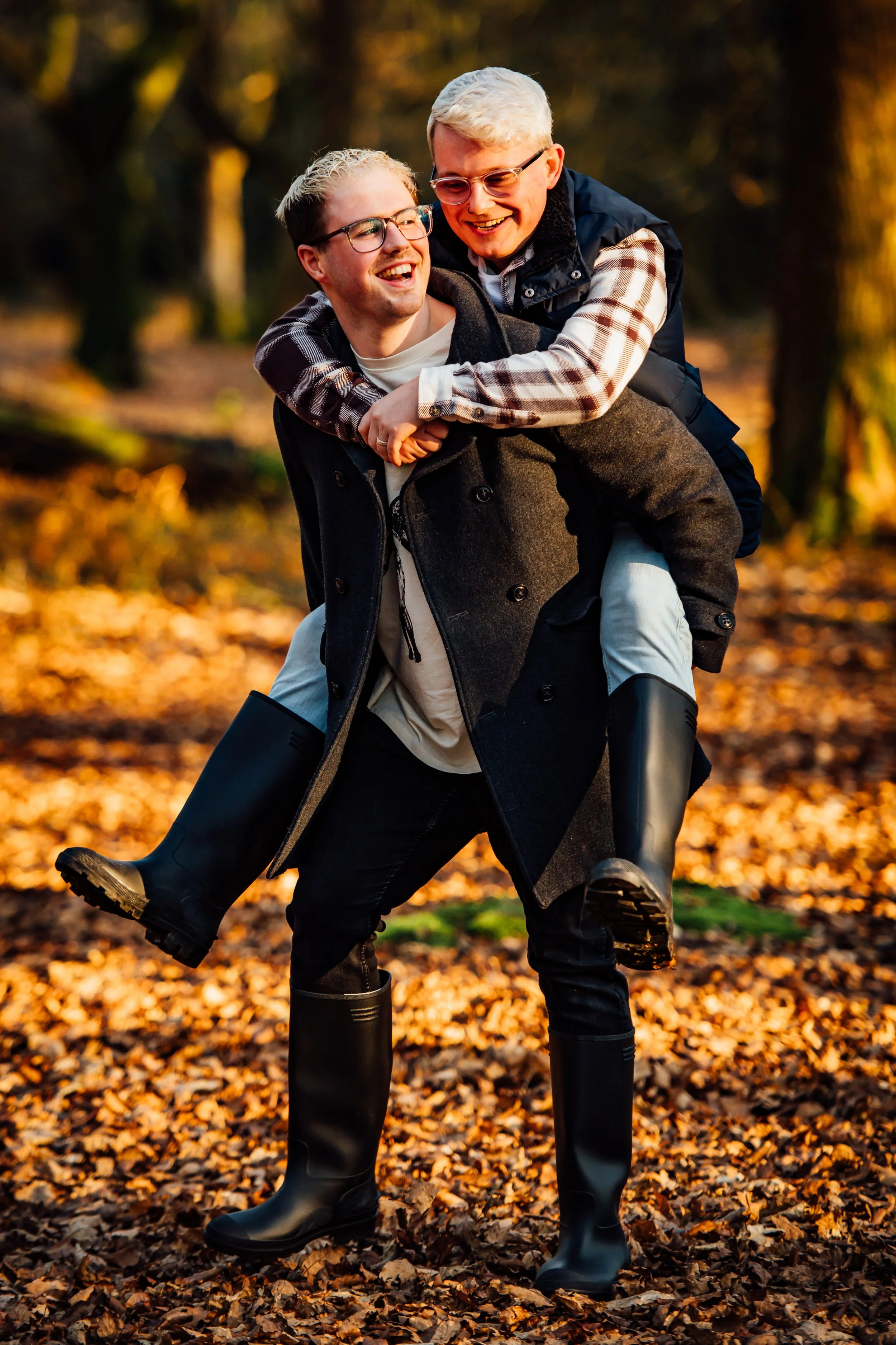 Two young men, wearing glasses and warm clothing, are outdoors in a forested area with autumn leaves on the ground. One man gives the other a piggyback ride, both smiling and enjoying the moment.