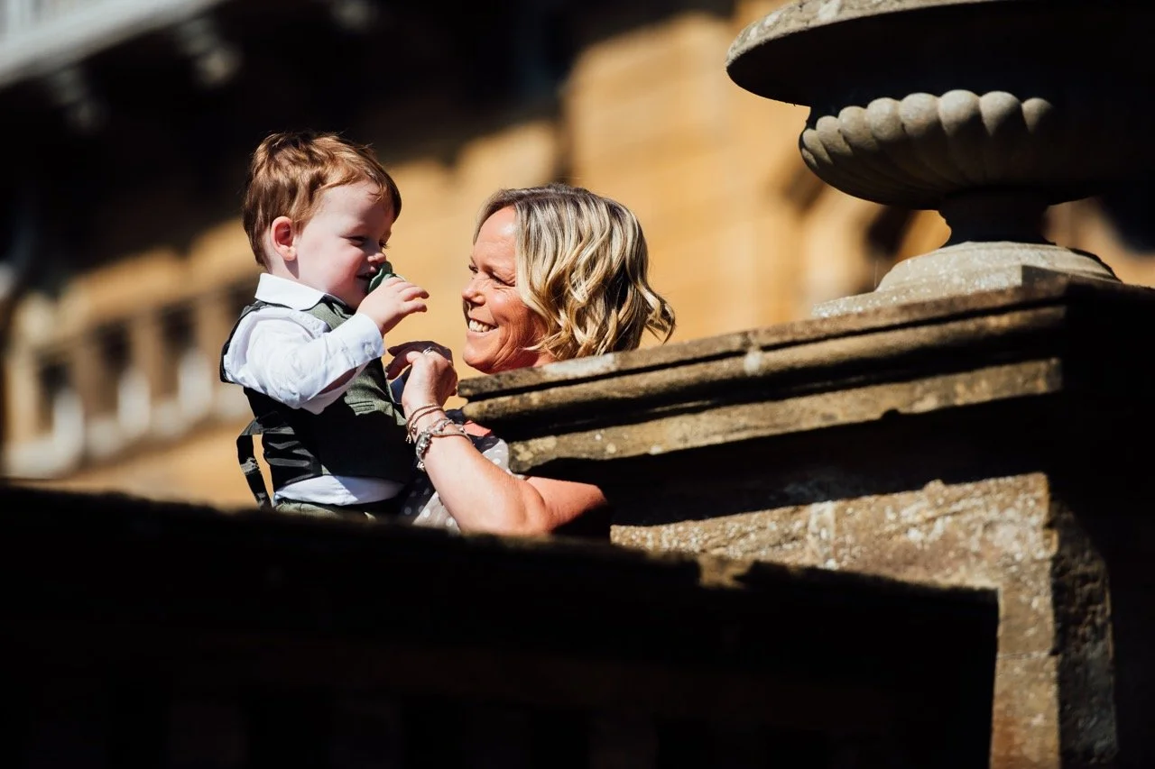 A woman smiling and holding a young boy with red hair, outdoors near a stone wall and a decorative stone fountain.
