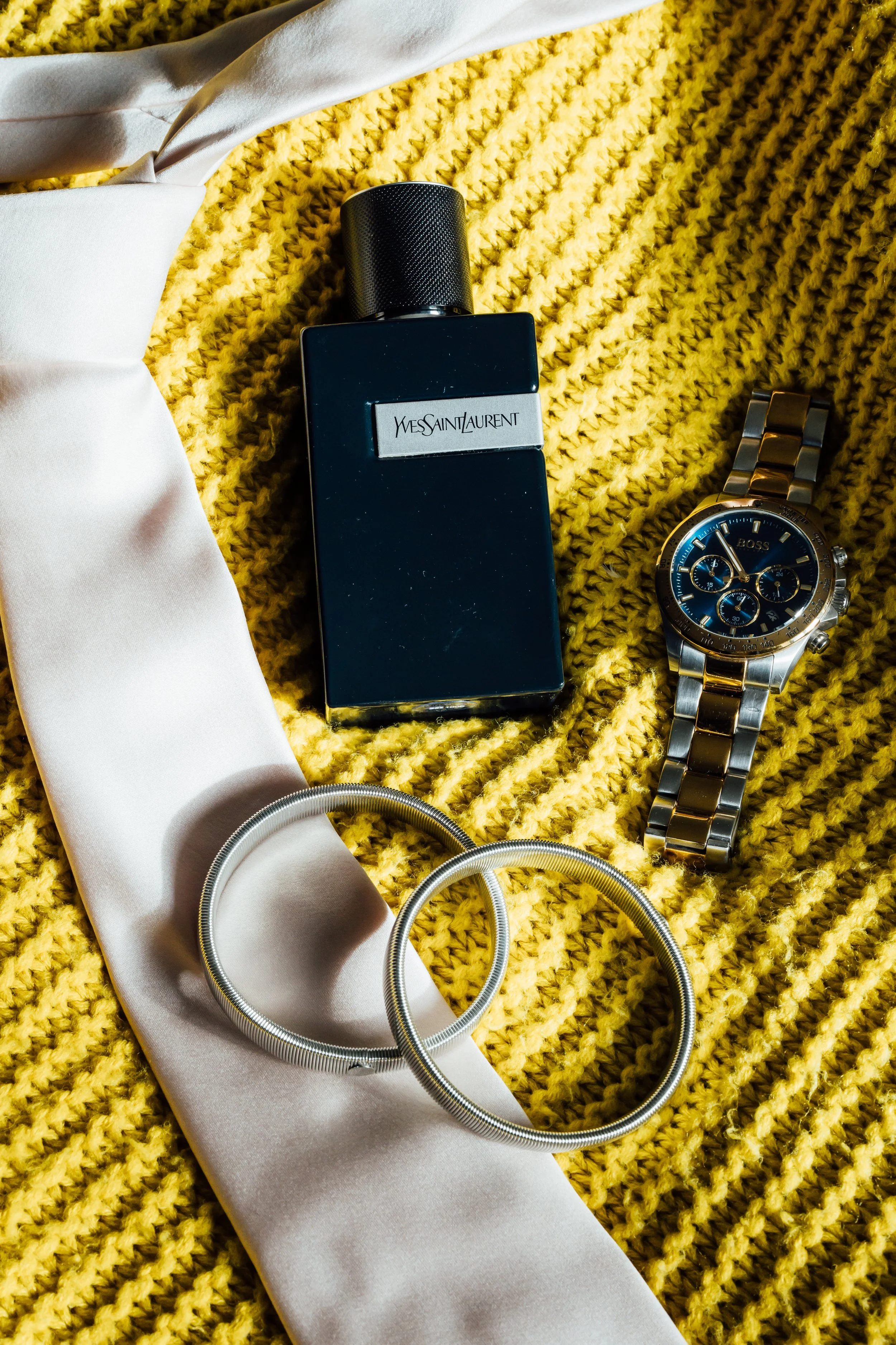 A perfume bottle, a wristwatch, a pair of silver hoop earrings, and a white silk scarf on a yellow textured fabric background.