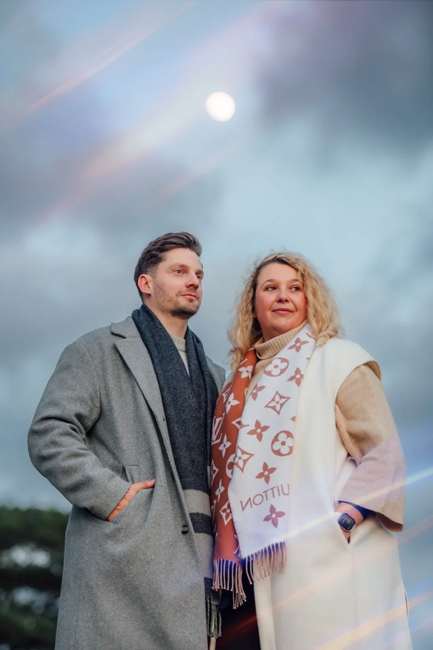 A man and woman stand outdoors under a cloudy sky with the moon visible, both dressed warmly and looking off into the distance.