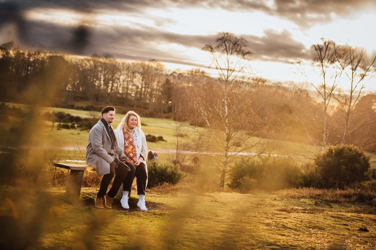 A couple sitting on a park bench during sunset, with trees and a grassy field in the background.