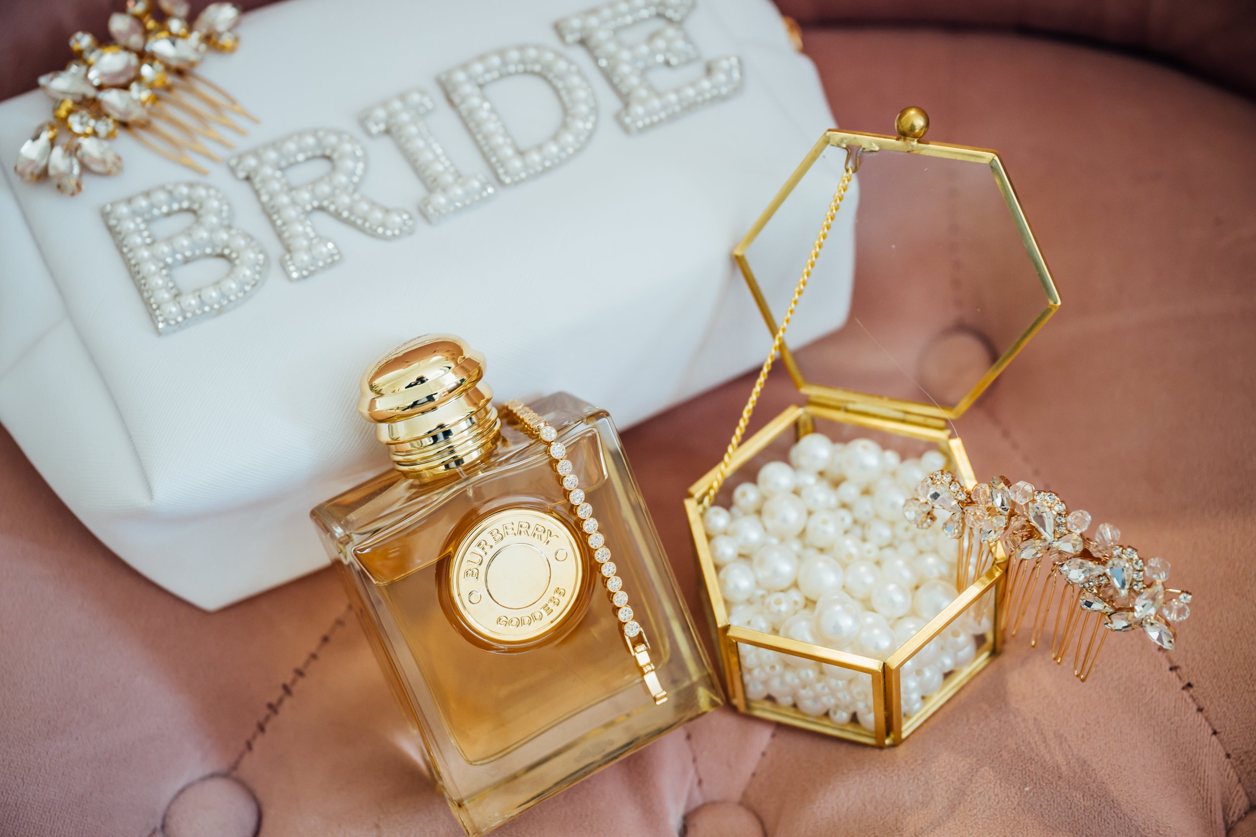 A bottle of perfume, pearl jewelry, and hair accessories on a pink fabric surface with a white box in the background.