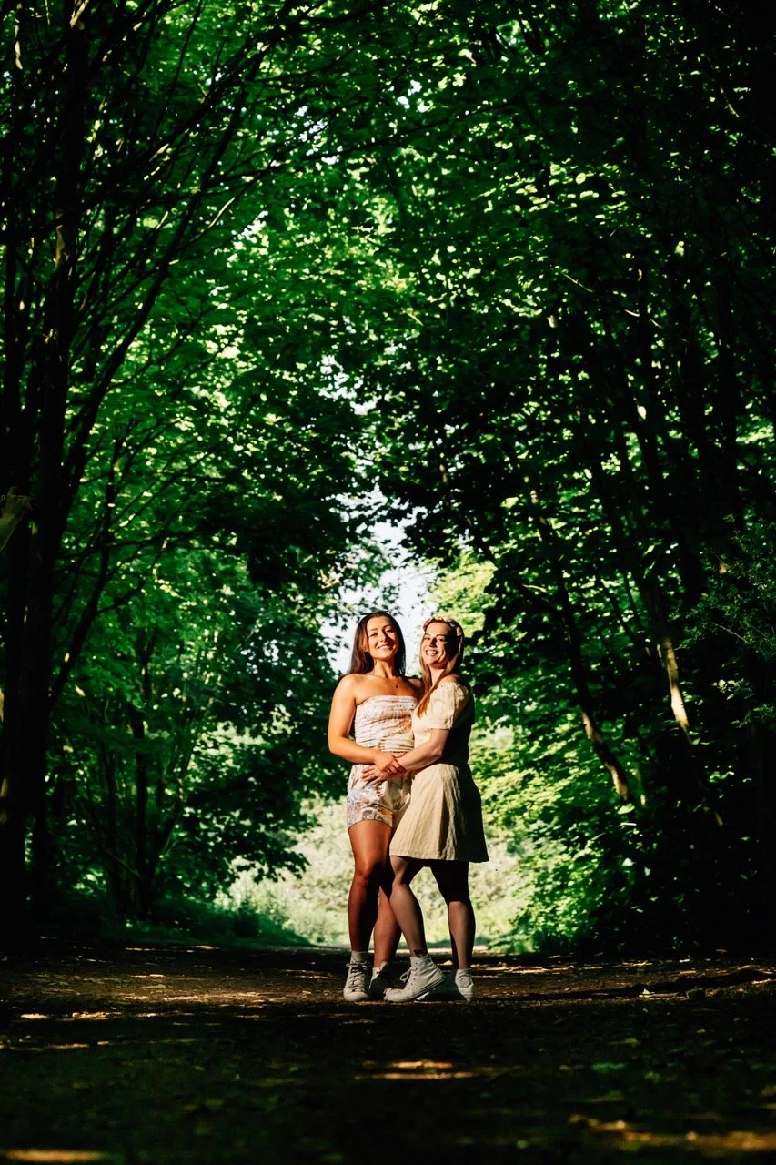 Two women standing together on a forest path, smiling, surrounded by lush green trees.
