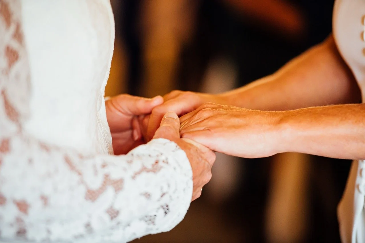 Close-up of two hands holding each other, one appears older with visible veins and wrinkles, and the other younger, in a white lace garment.