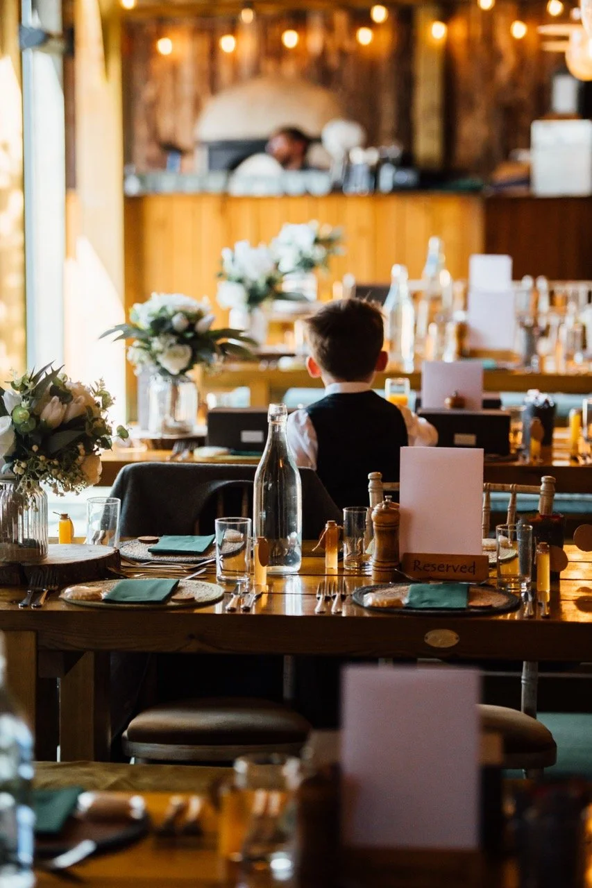 A dining room with tables set for a meal, decorated with floral centerpieces, and a boy sitting at a table facing away from the camera; a reserved sign is on one of the tables, and warm lighting creates a cozy atmosphere.