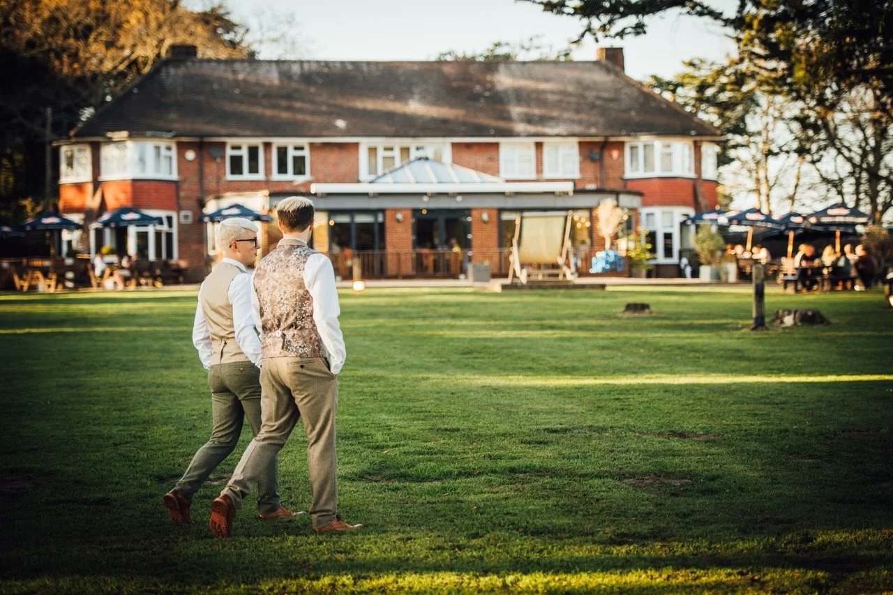 Two men walking on a grassy area near a large brick building with outdoor seating and umbrellas, during sunset.