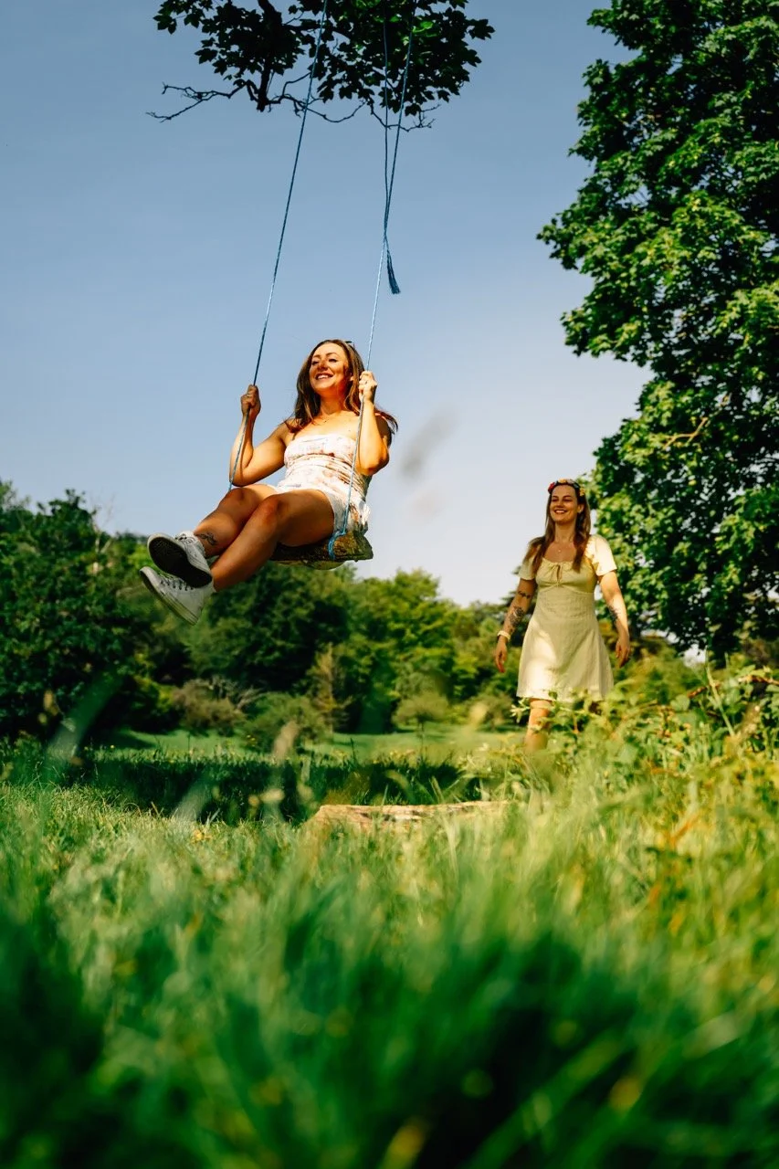 A woman with a floral headband in a light dress walks through a grassy park as another woman swings on a swing set, smiling under a clear blue sky surrounded by green trees.